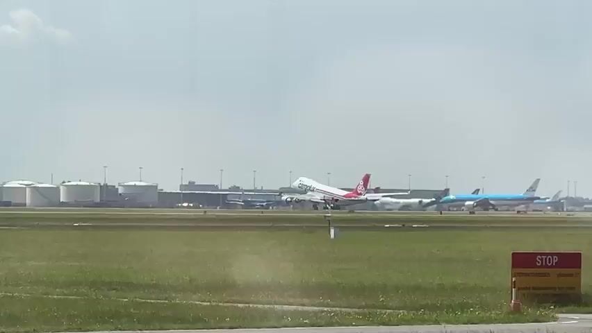 A passenger airplane with red and white livery is seen taking off on a cloudy day. The plane is ascending from a runway, with its landing gear retracted. In the background, there are several other airplanes parked on the tarmac, as well as large industrial storage tanks and a control tower. The ground in the foreground is covered with green grass. A red "STOP" sign is visible in the lower right corner of the frame.