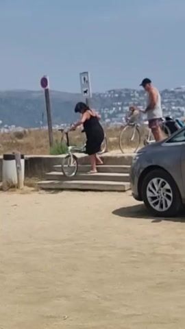 A woman in a black dress is pushing a bicycle up a set of concrete stairs. Her head is down and she appears to be concentrating on maneuvering the bike. Behind her, to the right, a man in a light-colored t-shirt and shorts is standing next to a bicycle with a dog in a basket on the front. He is looking towards the woman. In the foreground, a silver car is parked on a sandy surface. The background shows a grassy embankment with some buildings and hills beyond. There are also signs on poles near the stairs. The sky is clear and the sun is shining.