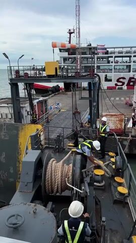 This video captures a scene at a ferry terminal in Tambon Don Sak, Thailand. The primary subjects are three individuals wearing safety vests and hard hats, actively engaged in securing a ferry. One person is operating a winch with a large coil of rope, while another is guiding the rope around bollards on the deck of a smaller vessel. A third person is positioned further back, observing the operation.

The setting is a busy port with a large ferry docked in the background, identifiable by its white hull and the "SEATRANS" logo prominently displayed. A ramp or gangway connects the ferry to the dock. The weather appears overcast, suggesting it might be morning or late afternoon. The sea is visible in the background.

The activity depicted is the docking or undocking of a vessel, specifically the process of securing it using ropes and a winch. The workers' attire and the presence of safety equipment indicate a professional maritime operation. The overall mood is one of focused work and efficiency, typical of a port environment. Visible text includes "SEATRANS" on the large ferry and "SEATRANS" on the safety vests of the workers. The windsock on the tall mast suggests moderate wind conditions.