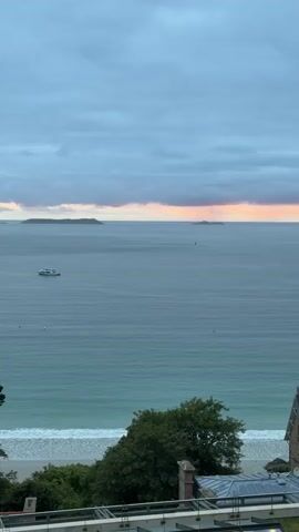 A panoramic view of the ocean at dusk. The sky is overcast with hues of pink and orange on the horizon, suggesting a sunset. In the distance, several small islands are visible. The sea is calm, with gentle waves breaking on a sandy beach in the foreground. There are a few boats on the water and some rocks jutting out from the sea. The foreground shows the roof of a building and some trees. The overall atmosphere is serene and tranquil.