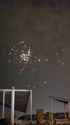 The video shows a night sky with a drone light show. Multiple drones are flying in formation, creating patterns of light against the dark sky. The drones are lit with various colors, including white, red, blue, and purple. Below the drones, there are the tops of some structures, possibly buildings or shelters, and a hint of a crowd of people. The drone formation appears to be in a spiral or circular pattern, with some drones forming a central cluster. The overall scene is one of a nighttime outdoor event.