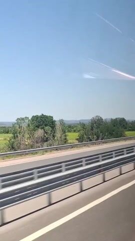 The video shows a highway scene. A silver car is driving on the highway in the distance. There are guardrails on the side of the highway. In the background, there are trees and green fields under a clear blue sky. There are some faint streaks in the sky, possibly contrails from airplanes. The camera appears to be moving, suggesting the video was filmed from a moving vehicle.