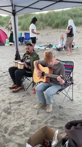 The video shows a group of people on a sandy beach, possibly at sunset, with dunes in the background. A white canopy provides shade. In the foreground, two young people are seated on folding chairs, playing musical instruments. One, a young person with red hair, is playing an acoustic guitar, wearing a grey t-shirt and blue jeans. Next to them, a man with dark hair and a beard is playing bongos, wearing a dark t-shirt, black pants, and a green cape. Behind them, several other people are visible. On the left, a person in a long green robe stands near a pink tent. In the middle ground, two people stand talking, one with long dark hair and the other wearing a white t-shirt. To the right, a person with long white hair stands with their back to the camera, and another person in a dark green patterned shirt with a circular emblem on the chest, possibly an amulet or medallion, is leaning forward, talking or gesturing. Another person in light-colored clothing is seated further back. A cardboard box with various items inside is visible in the bottom right corner of the frame. The scene appears casual and relaxed, with a potential gathering or performance taking place on the beach.