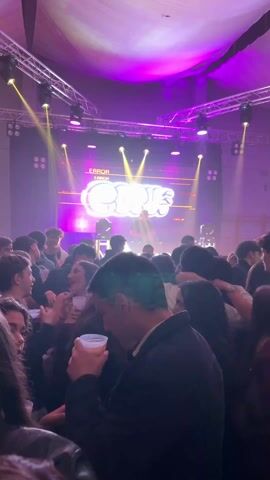 The video shows a lively party atmosphere.  A crowd of people are gathered, seemingly enjoying themselves, with drinks in hand. In the background is a stage with a DJ performing. The stage is lit with various colored lights, including yellow, purple, and blue.  A screen behind the DJ displays a graphic reading "ERROR". Someone in the crowd gives a thumbs up to the camera, then raises their hand with all fingers displayed.  The environment is indoors, with a visible ceiling structure and truss.