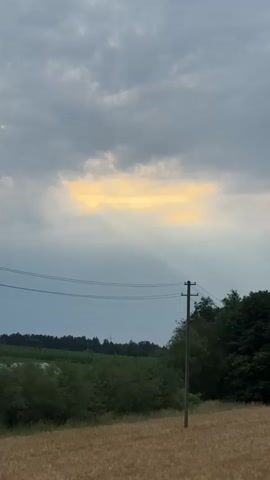 A short video shows a stormy sky with rays of sunlight breaking through the clouds over a rural landscape. In the foreground, a field of grain is visible, with a wooden utility pole and power lines running across the frame. In the distance, trees and vegetation line the horizon. A bird flies across the screen in the second and third clips.