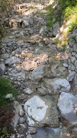 This is a high-angle, handheld video of a small mountain stream. The stream flows down a rocky bed, with stone walls lining both sides of the channel. The water is clear and shallow, running over smooth, grey rocks and some larger, white boulders. The surrounding environment appears to be forested, with greenery visible on the slopes above the stream. Sunlight filters through the trees, casting dappled shadows on the water and rocks. There are no people or animals visible in the video.