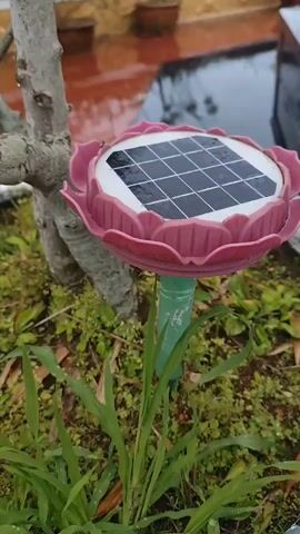 A close-up, slightly shaky video shows a pink, lotus-shaped solar-powered garden light. The light is positioned in a pot with some greenery around its base. The solar panel at the center of the "lotus" appears to be dusty. In the background, there is a reddish-orange tiled surface, possibly a patio, with two potted plants visible. The overall impression is that of an outdoor setting, possibly a garden or balcony, with the lighting device being the central focus.