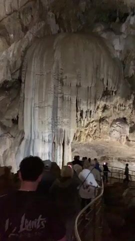 The video shows a group of people walking through a cave. The cave has large rock formations, including stalactites and stalagmites. The people are walking on a path that is lit by lights. Some people are taking pictures with their phones. The cave ceiling is rocky and uneven. The lighting in the cave is dim. There is music playing in the background.