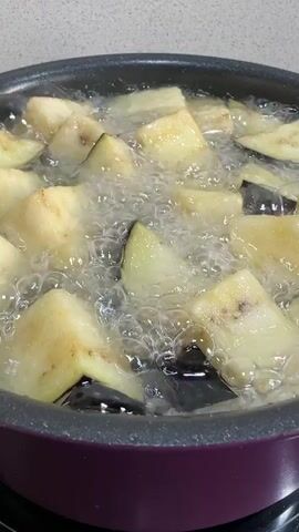 Chopped eggplant pieces are deep-frying in hot oil in a dark purple pot. The oil is bubbling vigorously, and the eggplant pieces are light yellow with some dark skin visible. The pot is on a stovetop, with a light-colored kitchen counter or wall in the background.