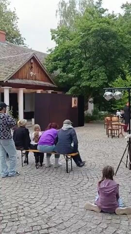 A small outdoor performance is taking place. In the foreground, a young girl with long brown hair, wearing a purple hooded sweatshirt and yellow leggings, sits on the ground facing away from the camera, her legs crossed. To her left, a group of people are seated on a wooden bench, facing the performance area. Among them are two women with long brown hair and a person wearing a blue jacket and a grey hood. Behind them, a wooden building with a brown roof is visible.

In the mid-ground, a makeshift stage is set up with a dark backdrop and a few chairs. Two performers are on the stage; one, wearing a black outfit with a white collar and black pants, stands to the left, while the other, dressed in a red jacket and dark pants, stands to the right and appears to be speaking. A lighting rig with multiple spotlights is positioned in front of the stage, and a speaker is set up to the right of the stage.

The audience is seated in chairs arranged in rows beyond the stage. They are a diverse group of adults and some children. A green tent is visible in the background, along with lush trees and foliage, suggesting a park or outdoor venue. The ground is paved with cobblestones. The overall atmosphere appears to be a casual, community-oriented event.