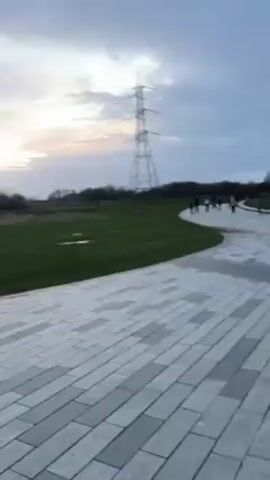 The video shows a park with two large, horse head sculptures. The sculptures are made of silver metal and are positioned facing opposite directions. People are walking around the sculptures and taking pictures. The park has a paved walkway and grassy areas. There is a power line tower in the background. The sky is cloudy with a hint of sunset.