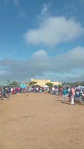 The video shows an outdoor gathering of people under a cloudy sky with a bright sun. The ground is dirt and appears to be a market or fair with various stalls and tents set up. In the foreground, a woman walks from right to left, wearing a dark green top and patterned shorts, carrying a white object. Several other people are visible in the mid-ground and background, some walking, some standing in groups. There are motorcycles parked on the right side of the scene. In the distance, there are buildings, including a light-colored, multi-story structure. The overall atmosphere seems to be one of casual assembly and activity.