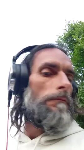 A close-up shot focuses on the face of a man with long, dark hair and a graying beard, wearing black headphones and a white hoodie. The man turns his head from left to right and directly at the camera, with his eyes seeming to stare. The background is blurred, showing hints of trees and an overcast sky.