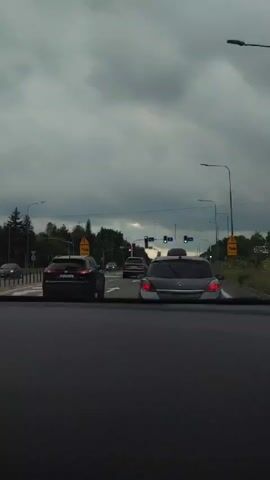 The video shows a view from inside a car, looking out through the windshield at a street scene. The sky is overcast and appears to be dusk or a cloudy day. In front of the car, there are other vehicles on the road. Traffic lights are visible, with some showing red and others green. Yellow warning signs are posted along the side of the road. The overall impression is of being in traffic, possibly at an intersection, with the weather contributing to a subdued atmosphere.