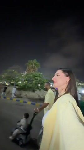A woman in a yellow top and white tank top is filming a video of herself while walking at night. In the background, a man is pushing a child in a small toy car. Other people are visible in the distance. The scene appears to be an outdoor urban area with streetlights and trees. The woman is smiling and looking at the camera.