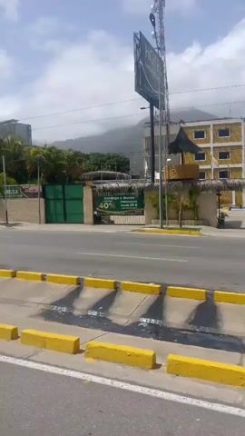 This video captures a dynamic street view in Caraballeda, Venezuela. The scene unfolds along a road bordered by yellow concrete curbs, suggesting a public thoroughfare. On the left side of the road, a hotel with a large sign is visible, featuring text that includes "HOTEL" and "40%." The hotel appears to be a significant structure, with other buildings and a communication tower in the background. Palm trees are scattered throughout the landscape, indicating a tropical or subtropical climate. The sky is partly cloudy, suggesting it is daytime with variable weather conditions.

The primary activity is the passage of vehicles. A white bus is seen traveling on the road, and other vehicles are implied by the road markings and infrastructure. The buildings in the background range from multi-story hotels or residential buildings to lower structures with white walls and blue accents. A prominent stone wall runs along some of the properties. In the distance, a mountainous landscape is visible, often characteristic of coastal regions. The overall impression is of a bustling urban or semi-urban environment in a warm climate. The presence of a hotel and the discount offer ("40%") suggest a location that caters to tourists or travelers. The time of day appears to be late morning or early afternoon, given the bright sunlight and shadows.