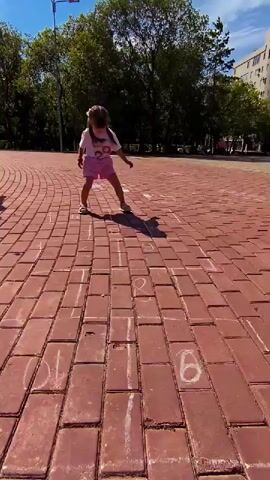 A young girl with long brown hair in pigtails, wearing a white t-shirt with cartoon characters and pink shorts, plays hopscotch on a red brick patio. The hopscotch grid is drawn with chalk, featuring numbers 1 through 10 in various squares. The girl is in mid-jump, with her shadow cast on the bricks. In the background, there are trees, a lamp post, and a residential building. The weather appears sunny and clear.