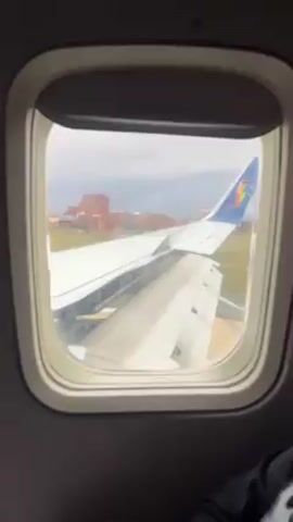 The video is taken from inside an airplane, looking out the window. A wing of the plane fills much of the window, with the tail visible in the distance. A rainbow-colored logo is on the tail. Beyond the wing, the scene shows an airport runway and some buildings in the background. The sky is overcast. A hand with a thumb raised appears briefly in the foreground, partially obscuring the view.