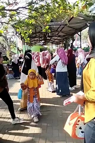 The video shows a group of children, dressed in traditional clothing, standing in a line outdoors. The children appear to be participating in an event or performance. Some adults are also present, supervising the children. The environment looks like a school or community gathering space, with buildings and trees visible in the background. The lighting suggests it is daytime, possibly sunny. The children are mostly looking towards the camera or in a consistent direction, indicating they may be waiting for instructions or to begin an activity. There are no obvious signs of distress or conflict.