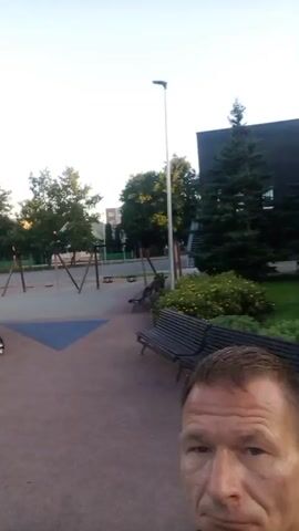 A man is recording a selfie video in a park with a playground. The playground features a large, castle-themed structure with slides and climbing elements. There are also benches and trees visible in the background. The man appears to be wearing earbuds and a dark shirt. The sun is low in the sky, suggesting it is either early morning or late evening.