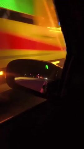 The video is filmed from the driver's perspective inside a car at night, with the dashboard and steering wheel visible in the foreground. The car is on a dark road, likely in the Baltezers area of Latvia.

Ahead, an emergency scene or convoy is visible, dominated by flashing blue and orange lights. Initially, an ambulance with its rear visible and prominent flashing lights is seen. Another vehicle, possibly police or fire services, also displays flashing lights further ahead. These lights cast a strong, pervasive blue glow over the road and surrounding area.

The car appears to be approaching or passing the emergency vehicles slowly. White road markings, including solid lines and directional arrows (chevrons), are visible on the asphalt. Small red and white road cones or markers are intermittently placed along the roadside. A blue circular traffic sign with a white arrow pointing right is visible on the right side of the road.

The time of day is clearly night, and the weather conditions appear clear. The scene conveys a sense of an active incident or response due to the presence and intensity of the emergency lights. No discernible text is visible on the vehicles or road signs within the video.