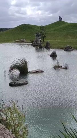 A wide shot captures a serene pond with ripples on its surface. Several large rocks are partially submerged in the water, with one featuring a clump of green reeds. In the background, rolling green hills are dotted with more rocks. On the crest of one hill, a small stone lantern stands near a lone tree. A group of four people is visible on the top of a distant hill, looking out at the landscape. The sky is overcast, with dramatic clouds. Green vegetation, including cattails, frames the bottom of the shot. The scene appears to be a park or garden, possibly with a Japanese aesthetic influence.