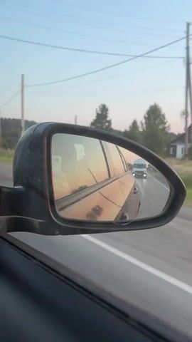 The video shows a view from inside a car looking out the side mirror. The side mirror reflects the road behind the car, where another vehicle, possibly a white van, is visible in the distance. The sky appears to be sunset or sunrise, with warm orange and pink hues reflected in the mirror and on the side of the car. Trees and power lines are visible along the side of the road in the background. The car is in motion, as indicated by the blurry road in the mirror and the slight motion blur of the surrounding environment.