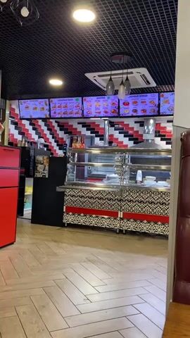 The video pans across the interior of a fast-food restaurant. The walls are decorated with a black, white, and red geometric pattern. Digital menu boards display food items above a service counter. The counter features a glass display case and stainless steel equipment. To the left, a red refrigerator and a black cabinet are visible. The flooring is light-colored wood laid in a herringbone pattern. A red leather booth is partially visible in the foreground on the right. The overall atmosphere appears clean and modern.