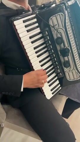 A person in a black suit and white shirt plays an accordion. The accordion is black and silver with white keys and black keys. The person's hands are on the keys, and their fingers are moving. The background is blurred, but it appears to be an indoor setting with some furniture. The video is in color and the lighting is adequate.