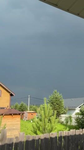 The video captures a view of a suburban or rural backyard under a darkening, overcast sky. The foreground features a weathered wooden fence. Beyond the fence, the scene includes a small, light brown wooden shed to the left, a taller yellow house with a brown roof to the far left, and a section of a white house with a grey roof to the right. A vibrant green pine sapling stands prominently in the center, with larger evergreen trees scattered throughout the mid-ground. A utility pole with wires is visible behind the sapling. The sky is a deep, stormy grey, suggesting an approaching storm or the aftermath of one. The bright green grass and the yellow house suggest it is daytime, likely late afternoon or early evening, as the light seems to be diminishing. The overall mood is somber and anticipatory due to the dark sky. No people are visible, and there is no discernible text in the frame. The location context provided is Бобровский, Russia.