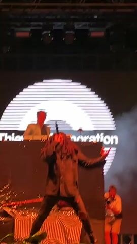 A live music performance is depicted with a lead singer in the foreground, a DJ in the background, and a cameraman to the right. The lead singer, with dreadlocks, is wearing a black jacket and ripped pants, kneeling on stage and singing into a microphone. The DJ, wearing glasses and a light-colored blazer, is behind a console. The cameraman, wearing a t-shirt and shorts, is filming the performance. The stage features a large circular backdrop with the words "Thieves Corporation" and several rows of horizontal lines. Lighting rigs with colorful lights are visible above the stage. Smoke is present in the air, and some band equipment, including a guitar, is on the left side of the stage. The audience is partially visible at the bottom of the frame.