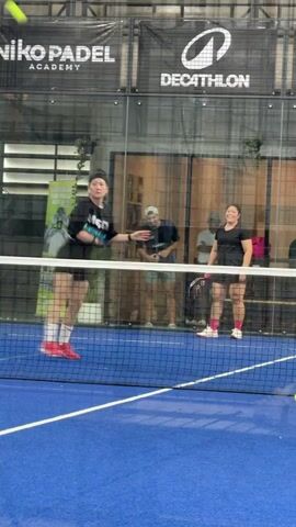 A young woman in a black t-shirt and black shorts with red and white sneakers is playing padel. She is on a blue court with a net in the foreground and glass walls behind her. She is mid-swing, hitting a ball with a padel racket. Her mouth is open, and she is smiling. In the background, behind the net, another woman in a black t-shirt and black shorts is also playing, and two men are watching. A sign above the court reads "NIKO PADEL ACADEMY" and "DECATHLON". The lighting is bright, suggesting an indoor court.