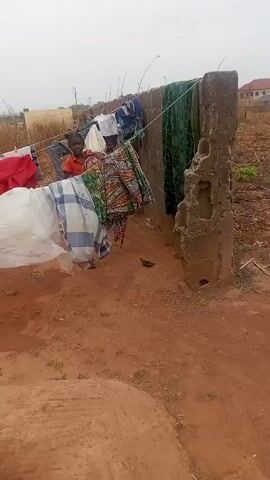 A young Black girl with her hair in two buns is standing behind a clothesline, smiling and interacting with someone who is filming. The clothesline is strung between a wall and a post, both made of concrete, and has a variety of clothing items hanging on it. The ground is dirt with some vegetation and trash scattered around. In the background, there is a field of dry grass. The weather appears overcast.