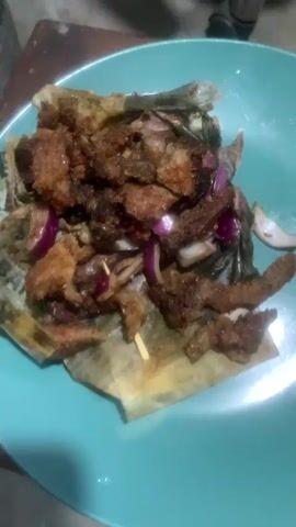 A close-up shot of a plate of food. The plate is turquoise and filled with a dish that appears to be fried meat and red onions. The food is served on a piece of paper that has writing on it. The background is out of focus and appears to be a table and a wall. The lighting is dim.