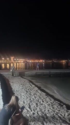 The video opens on a dark night, focusing on a sandy beach in the foreground. A person, partially visible from behind, sits on the sand, looking at their phone. To the left, a woman in a patterned dress walks along the beach. In the middle ground, a walkway or pier extends over the water. Several people are visible on the pier, some walking and one man standing. Lights from a distant town or resort illuminate the background, reflecting on the calm water. A Ferris wheel, lit with colorful lights, is visible in the distance and appears to change colors from red to green during the video. The scene depicts a lively waterfront at night.