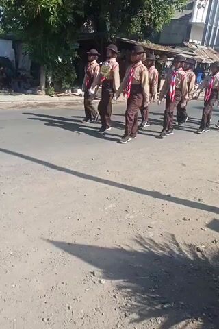 A group of young people in scout uniforms are marching in formation along a dirt road. The scouts are wearing khaki shirts, brown pants, and brown hats. They are also carrying red and white scarves. The formation is in two rows of four scouts each. They are marching in step and appear to be in a parade or a march. The sun is shining brightly, and there are shadows on the ground. There are trees and buildings in the background.