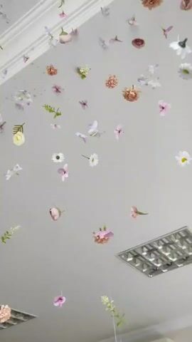 The video shows a ceiling decorated with numerous artificial flowers of various colors and sizes suspended by thin strings. The flowers are scattered across the ceiling in a seemingly random arrangement, creating a whimsical and ethereal effect. Some flowers are close to the ceiling, while others hang lower. There are also what appear to be air vents or light fixtures visible on the ceiling. The lighting is soft and diffused. There are no people or animals visible in the video. The overall impression is one of delicate beauty and lightness.