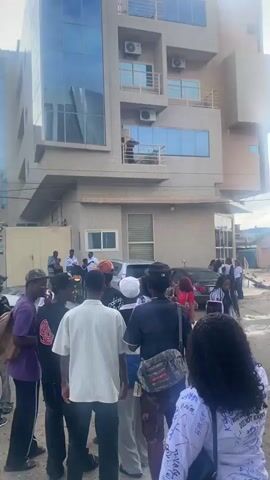 The video shows a group of people gathered outside a multi-story building. Many individuals are wearing white shirts, some with signatures or messages written on them. There are several cars parked in the vicinity, including a silver sedan, a black sedan, and a white SUV. Some people appear to be interacting, with gestures and conversations. The environment is an outdoor urban setting with a paved or dirt surface. The building behind the group has multiple floors with windows and air conditioning units. The sky is partly cloudy. The video pans and shifts focus, capturing different angles of the scene and individuals.