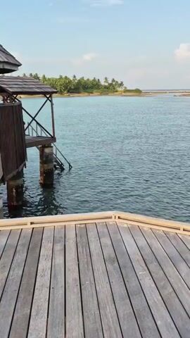 A short video shows a pier extending over shallow turquoise water. The pier is made of light-colored wooden planks. A set of stairs with a metal railing leads down into the water. The water is clear enough to see the bottom, which is covered in sand and some dark rocks. In the distance is a low-lying island with palm trees. The sky is pale blue with some wispy white clouds.