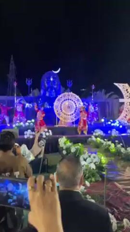 The video depicts a nighttime outdoor celebration featuring a stage with a Shiva head statue illuminated in blue, a large white-and-gold circular decoration, and a crescent moon decoration to the right of the stage. Five dancers wearing red outfits are performing on the stage. The foreground shows several people in the audience; some of them are holding their phones to record the performance. White flower arrangements adorn the front of the stage area. The overall setting suggests a wedding or similar festive event.
