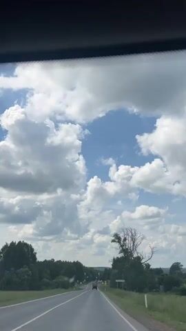 This is a first-person perspective video recorded from inside a vehicle traveling on a paved road. The road stretches into the distance, with trees lining both sides. The sky is filled with dramatic white clouds against a bright blue backdrop. A small truck is visible in the distance on the road. There is a road sign on the right side of the road. The video captures a serene rural landscape under a cloudy sky.