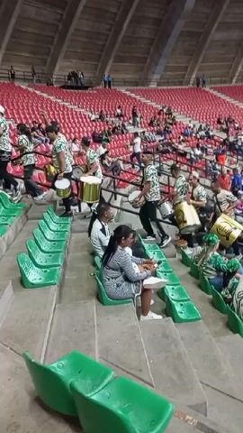 The video captures a scene inside a large stadium with predominantly red seating. In the foreground, a group of individuals are playing large drums, moving energetically through the rows of green seats. Some people are seated, watching the drummers or occupied with their phones. The drummers, mostly men, are wearing matching outfits, while the seated spectators are dressed in a variety of casual clothing. The stadium has a high, vaulted ceiling, and other spectators can be seen scattered throughout the stadium in the background.