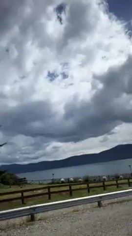 The video shows a view from a vehicle moving along a road beside a body of water, possibly a lake or sea.  Across the water, hills or mountains are visible in the distance. The sky is filled with dramatic, heavy clouds, though some patches of blue are visible. Beside the road, there is a grassy area with a wooden fence, a guardrail, and a few trees. As the vehicle progresses, a large building with a red roof becomes visible. The building appears to be a hotel or lodge. A smaller, stone building is visible nearby. Several cars are parked outside the main building. Power lines run above the road.