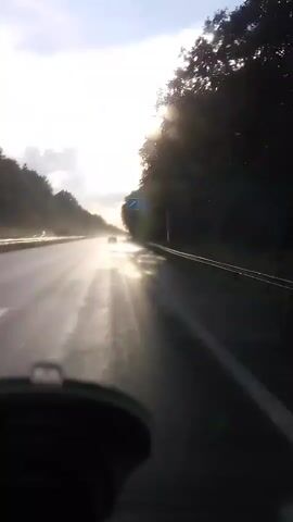 The video is filmed from the perspective of a driver on a wet road, likely a highway, with trees lining both sides. The sun is low in the sky, creating a bright glare and reflecting off the wet asphalt, making it difficult to see far ahead. A single car is visible in the distance on the same road, appearing as a dark silhouette against the bright sun. The camera appears to be mounted on the dashboard or windshield, as the bottom of a black object, possibly a dashcam or phone mount, is visible in the foreground. The sky is partly cloudy, with streaks of sunlight breaking through. There is a sense of movement as the scenery passes by.