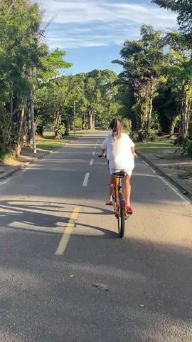 A young woman with long brown hair is riding a bicycle on a paved path lined with trees. She is wearing a white t-shirt and dark shorts. The path has yellow lines painted on it. The sun is shining and the sky is blue with some clouds. The trees are lush and green. The scene appears to be a park or a recreational area.