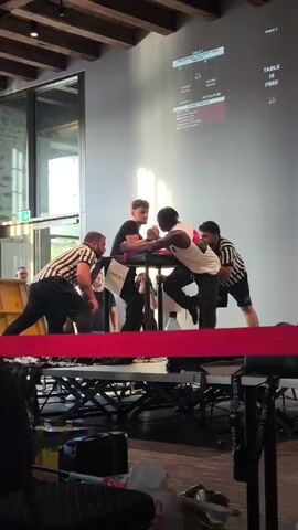 A video captures an arm wrestling match in progress. Two individuals, one appearing to be of lighter complexion and the other of darker complexion, are locked in a struggle, their arms positioned on a padded table. Two referees, dressed in black and white striped shirts, stand on either side of the competitors, observing the match closely. The environment appears to be an indoor venue with wooden beams visible in the ceiling and large windows in the background. A large screen displays graphics that seem to be related to the competition, including text indicating "TABLE IS FREE." In the foreground, a red ribbon is stretched across a raised platform. The lighting suggests it may be daytime, with sunlight streaming through the windows. Several other people are visible in the background, some observing the match, others seemingly involved in the event organization. The overall scene depicts a competitive sporting event focused on arm wrestling.