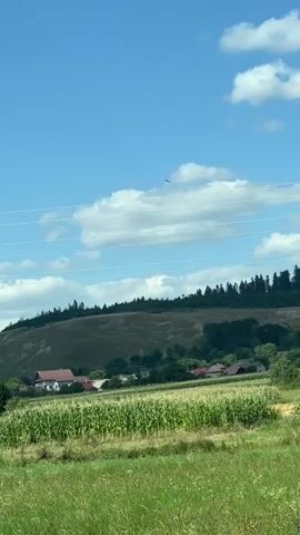 The video shows a rural landscape on a bright, sunny day. The sky is a vibrant blue with scattered white clouds. In the foreground, there is a lush green field. Beyond this field, a cornfield is visible, with rows of tall green stalks. In the mid-ground, several houses with red roofs are nestled among trees and rolling hills. A dense line of dark green trees forms the crest of a hill in the background. The overall impression is one of a peaceful, idyllic countryside.