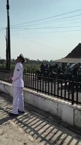 A group of people in white uniforms are gathered outside. One person in the foreground is looking up at a pole. In the background, other people in white uniforms are standing behind a fence. There is a flag on a pole in the distance. The ground is paved, and there are shadows on the ground. The sky is clear and blue.