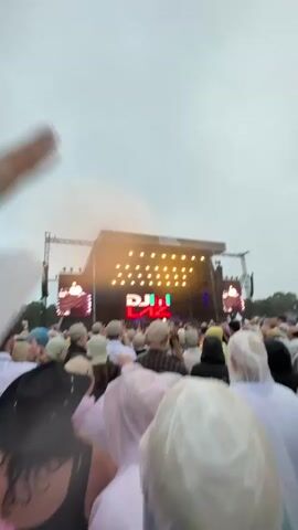 A crowd of people, many wearing rain ponchos, stands in front of a large outdoor stage with a screen displaying "DJI" in large red letters. The stage is lit with numerous yellow lights. The sky is overcast and appears to be raining. Some people in the crowd are raising their hands. A person's hand is visible in the upper left corner of the frame, reaching out. The video appears to be shot from a low angle, looking up at the stage.