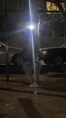A person is sitting on a swing in a dimly lit outdoor area at night. They are wearing light-colored pants and a dark top. The swing set has a metal frame, and the person is holding onto chains. The background shows a building with lit windows and two cars parked nearby, one silver and one black. The lighting is harsh, with bright spotlights creating lens flares. The ground is covered in dirt and debris.